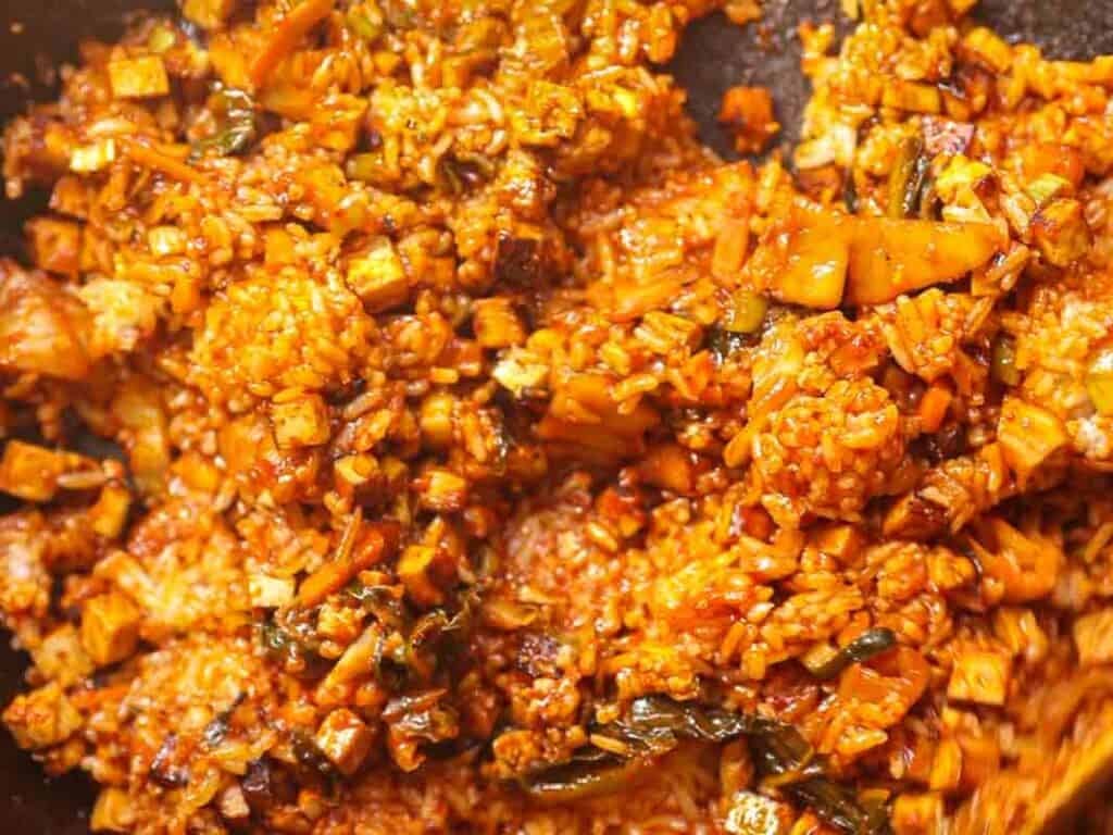Close-up of spicy kimchi fried rice in a pan, featuring cooked rice mixed with kimchi, vegetables, and seasonings. The dish appears vibrant and well-cooked, with a reddish hue from the spices.